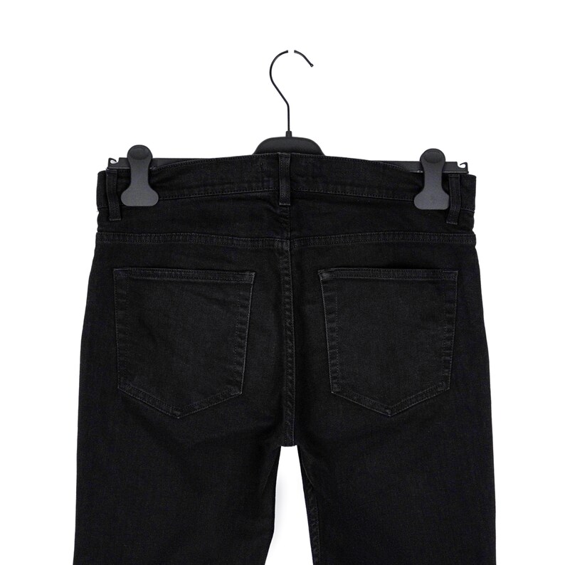 May include: Black denim jeans hanging on a black hanger. The jeans have two back pockets and a classic five-pocket design. The garment is made of a dark, solid-colored fabric.