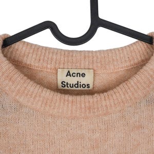Pu&ograve; includere: Un maglione color pesca chiaro con scollo tondo, appeso a una gruccia nera. L'etichetta recita "Acne Studios". La maglia ha un aspetto morbido e strutturato. Il maglione &egrave; un capo d'abbigliamento classico.