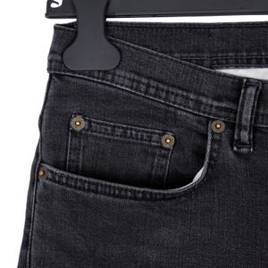 May include: Close-up of black denim jeans with a classic five-pocket design. The jeans feature brass-colored rivets and a button closure. The fabric has a slightly faded appearance, and the jeans are hanging on a black hanger.