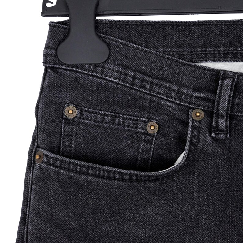 May include: Close-up of black denim jeans with a classic five-pocket design. The jeans feature brass-colored rivets and a button closure. The fabric has a slightly faded appearance, and the jeans are hanging on a black hanger.