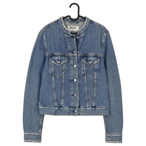 Op de afbeelding: Een lichtblauwe spijkerjas met een klassiek design, met een ronde kraag, knoopsluiting en twee borstzakken. De jas hangt aan een zwarte hanger. Het label luidt "Acne Studios".