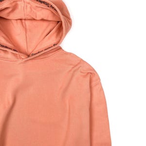 May include: A peach-colored hoodie with a drawstring hood. The hood features the text "Acne Studios" in black. The bottom hem of the hoodie has a distressed, frayed edge. The hoodie is made of a soft, comfortable material.