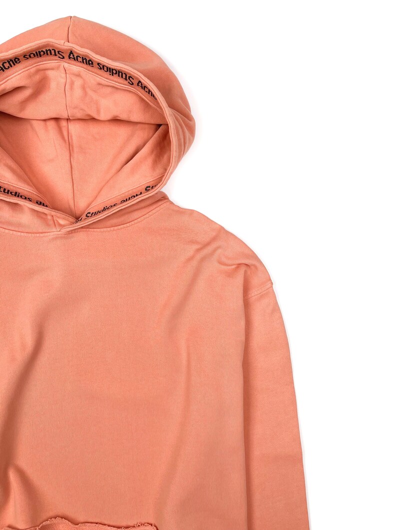 May include: A peach-colored hoodie with a drawstring hood. The hood features the text "Acne Studios" in black. The bottom hem of the hoodie has a distressed, frayed edge. The hoodie is made of a soft, comfortable material.