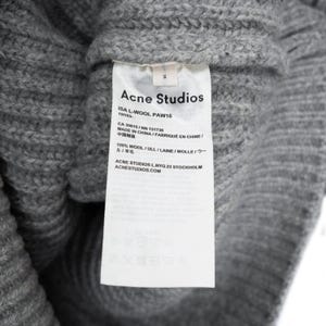 以下が含まれることがあります： グレーのニットウェアのクローズアップ。白いタグには「Acne Studios」と記載されており、素材の詳細（ウール100％など）が含まれています。衣類には「S」と表示されています。
