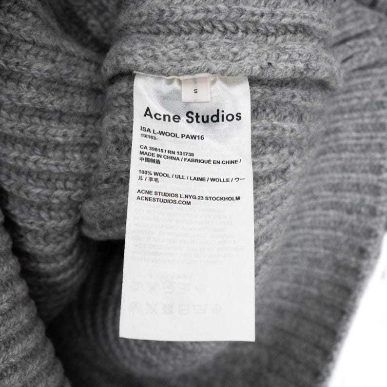 以下が含まれることがあります： グレーのニットウェアのクローズアップ。白いタグには「Acne Studios」と記載されており、素材の詳細（ウール100％など）が含まれています。衣類には「S」と表示されています。