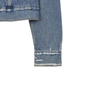 Op de afbeelding: Detail van een lichtblauwe spijkerjas mouw. De jas heeft een versleten uitstraling met gerafelde randen aan de manchet. De denim stof vertoont subtiele kleurvariaties, typerend voor een gedragen kledingstuk.
