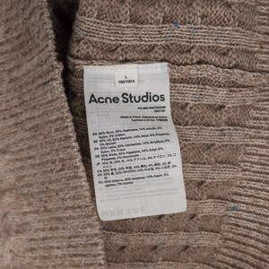 May include: Close-up of a beige cable-knit sweater with a tag from Acne Studios. The tag lists the materials: wool, cashmere, acrylic, nylon, and cotton. The sweater is size L, 185/100A.