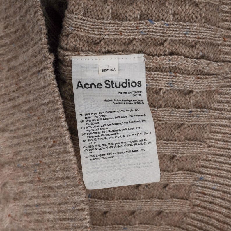 May include: Close-up of a beige cable-knit sweater with a tag from Acne Studios. The tag lists the materials: wool, cashmere, acrylic, nylon, and cotton. The sweater is size L, 185/100A.