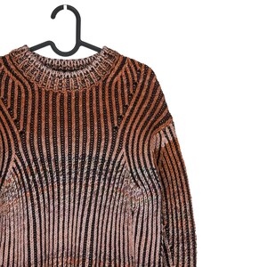 May include: A striped sweater in shades of brown, black, and pink hangs on a black hanger. The sweater features a ribbed knit design with vertical stripes. The neckline and shoulders are visible, showcasing the garment's texture and pattern.