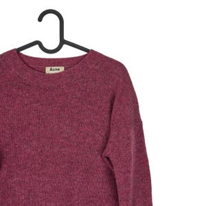 May include: A burgundy crew neck sweater hanging on a black hanger. The sweater has a textured knit and a small label that reads "Acne". The garment is made of a soft material.