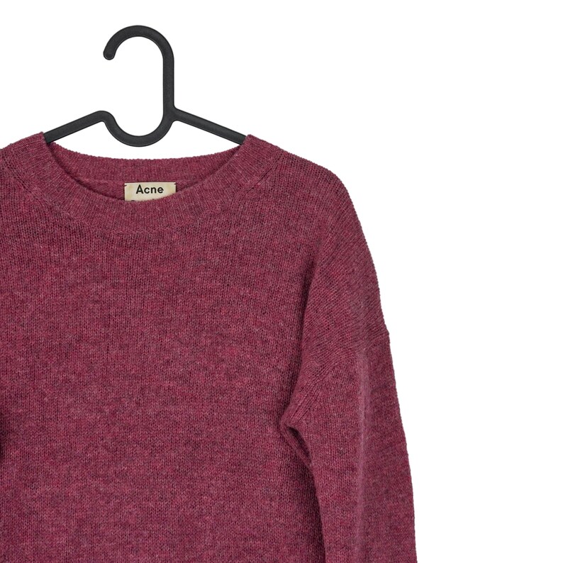 May include: A burgundy crew neck sweater hanging on a black hanger. The sweater has a textured knit and a small label that reads "Acne". The garment is made of a soft material.