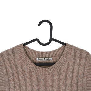 May include: A light brown cable knit sweater by Acne Studios hangs on a black hanger. The sweater has a crew neck and a textured, woven design. The brand's label is visible on the neckline.