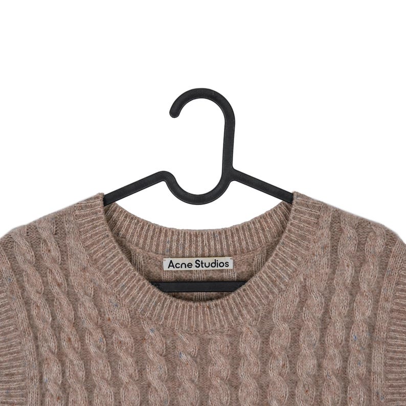 May include: A light brown cable knit sweater by Acne Studios hangs on a black hanger. The sweater has a crew neck and a textured, woven design. The brand's label is visible on the neckline.