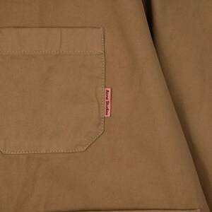 May include: Close-up of a tan-colored garment with a square pocket on the left side. A small pink tag with the text "Acne Studios" is sewn on the right side of the garment. The fabric appears to be a sturdy cotton or similar material.