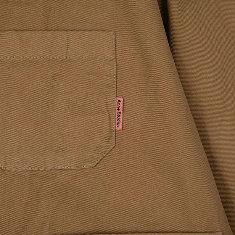May include: Close-up of a tan-colored garment with a square pocket on the left side. A small pink tag with the text "Acne Studios" is sewn on the right side of the garment. The fabric appears to be a sturdy cotton or similar material.
