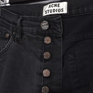 Pu&ograve; includere: Primo piano di jeans neri in denim con chiusura a bottoni color argento. I jeans presentano l'etichetta "ACNE STUDIOS". I bottoni sono impressi con il nome del marchio. I jeans sono di un lavaggio scuro.