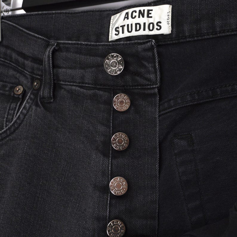 Pu&ograve; includere: Primo piano di jeans neri in denim con chiusura a bottoni color argento. I jeans presentano l'etichetta "ACNE STUDIOS". I bottoni sono impressi con il nome del marchio. I jeans sono di un lavaggio scuro.