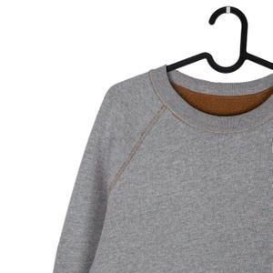 May include: A grey sweatshirt with a brown lining hangs on a black hanger. The crew neck sweatshirt has raglan sleeves with visible stitching. The fabric appears to be a soft, comfortable material.