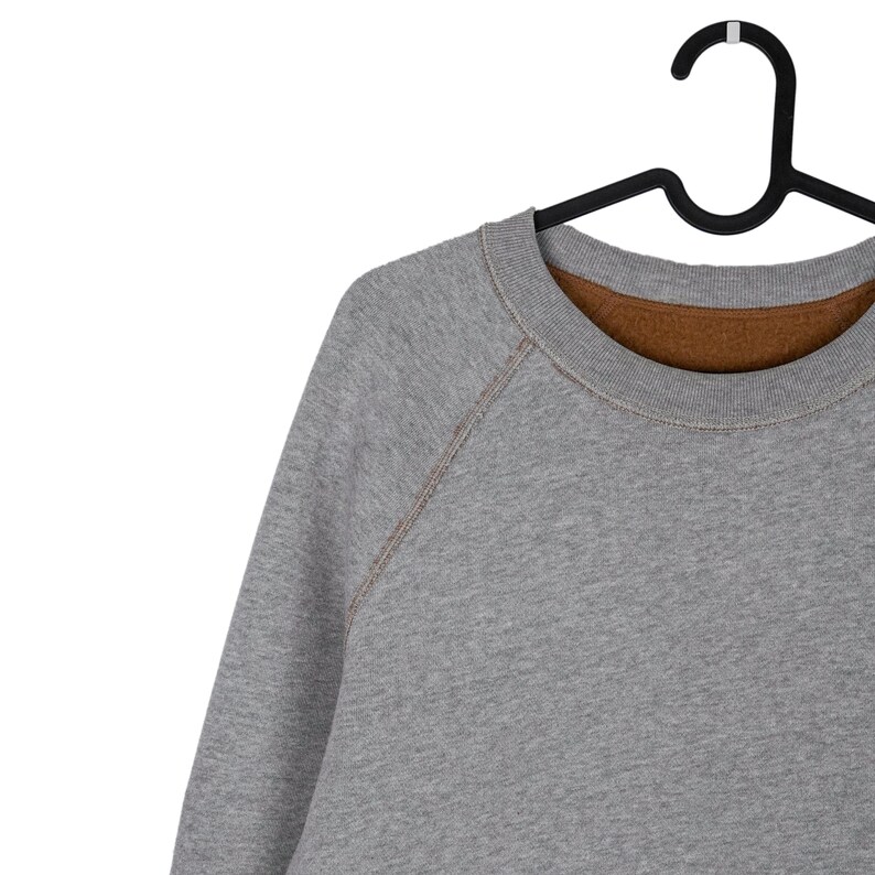 May include: A grey sweatshirt with a brown lining hangs on a black hanger. The crew neck sweatshirt has raglan sleeves with visible stitching. The fabric appears to be a soft, comfortable material.