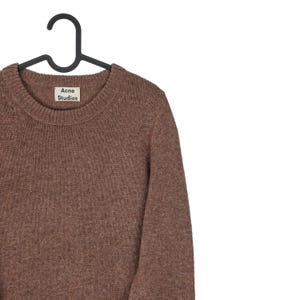 Peut inclure: Un pull marron &agrave; col rond suspendu &agrave; un cintre noir. Le pull a un aspect textur&eacute; et une petite &eacute;tiquette qui indique "Acne Studios". Le pull est fait d'une mati&egrave;re douce.