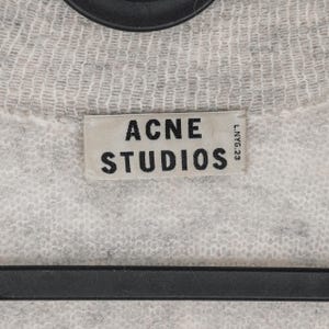 Puede incluir: Primer plano de un su&eacute;ter de punto gris claro con la etiqueta "ACNE STUDIOS" en un lugar destacado. La etiqueta rectangular tiene texto negro sobre un fondo gris claro. El su&eacute;ter tiene una apariencia texturizada.