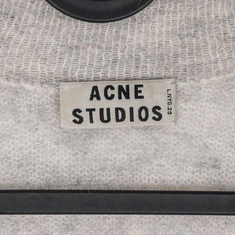 Puede incluir: Primer plano de un su&eacute;ter de punto gris claro con la etiqueta "ACNE STUDIOS" en un lugar destacado. La etiqueta rectangular tiene texto negro sobre un fondo gris claro. El su&eacute;ter tiene una apariencia texturizada.