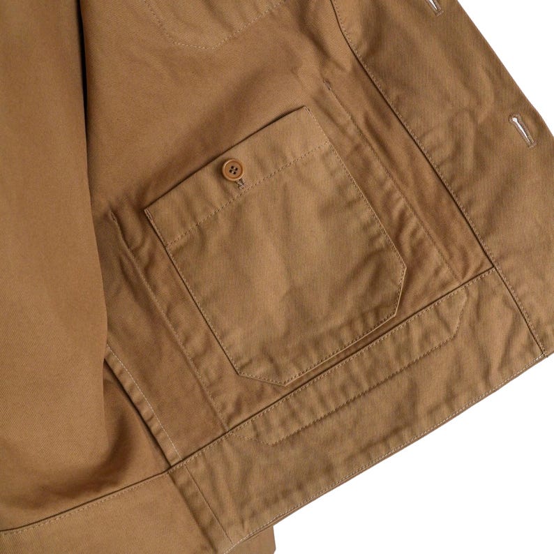May include: Close-up of a tan jacket with a pocket and a button. The jacket is made of a sturdy fabric and has visible stitching. The pocket is rectangular and has a button closure. The overall design is simple and functional.