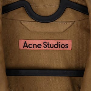 May include: Close-up of a tan jacket with the text "Acne Studios" on a pink label. The jacket is hanging on a black hanger. The fabric appears to be a sturdy cotton or similar material.