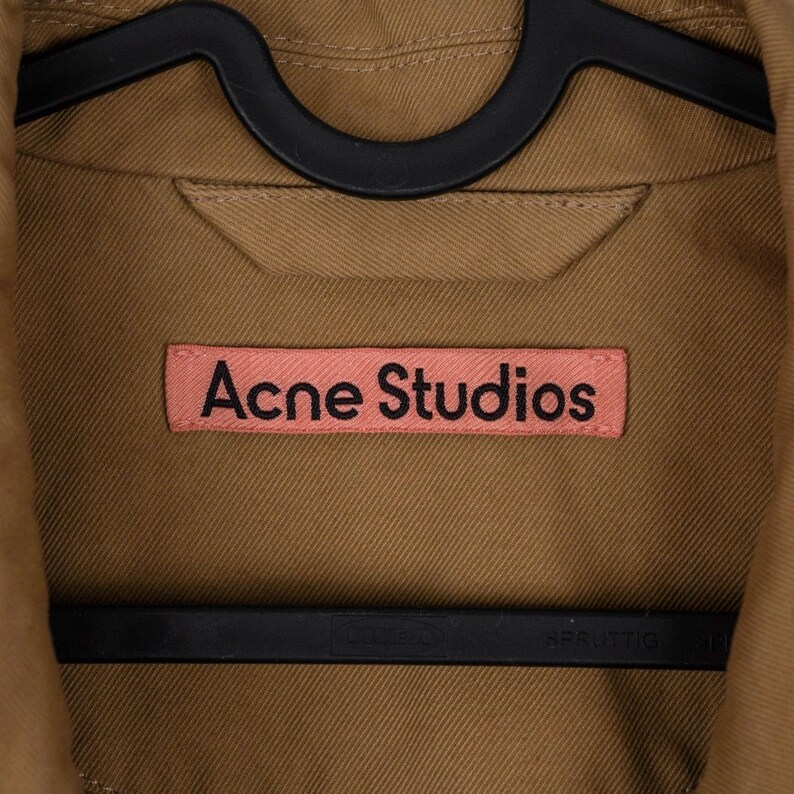 May include: Close-up of a tan jacket with the text "Acne Studios" on a pink label. The jacket is hanging on a black hanger. The fabric appears to be a sturdy cotton or similar material.