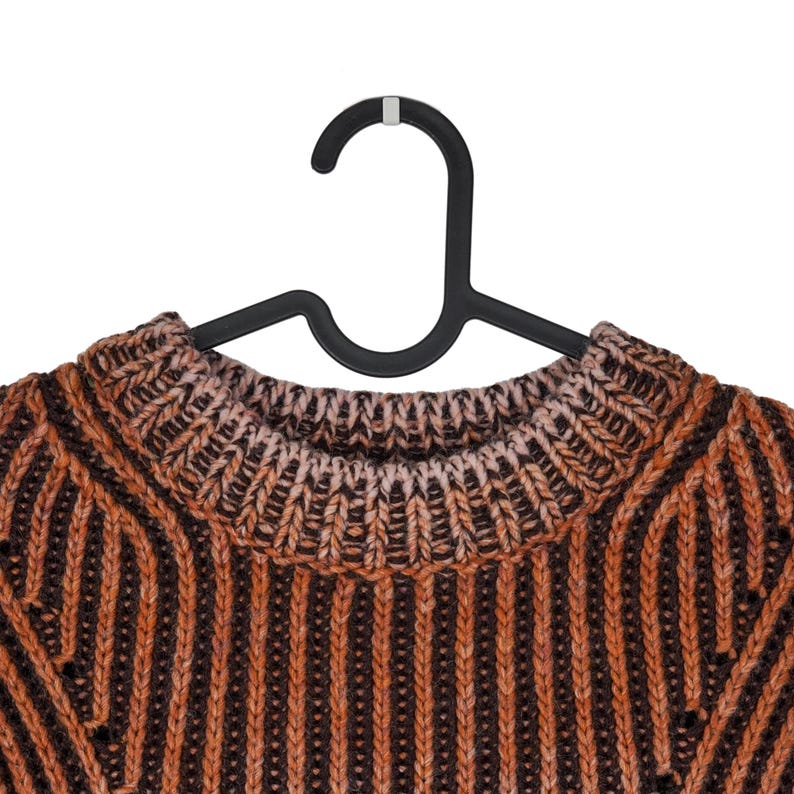May include: Close-up of a brown and orange knit sweater on a black hanger. The sweater has a ribbed pattern with vertical stripes. The neckline is a lighter shade of brown.