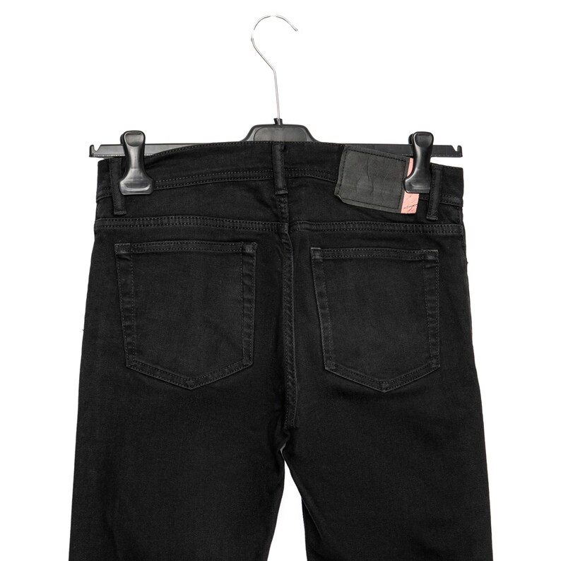 May include: Black denim jeans hanging on a black hanger. The jeans feature two back pockets and a leather patch. The denim is a solid black colour.