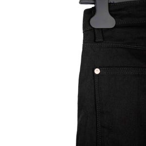 May include: Close-up of black denim jeans hanging on a black hanger. The jeans have a classic five-pocket design with a silver button on the back pocket. The stitching is a matching black color.