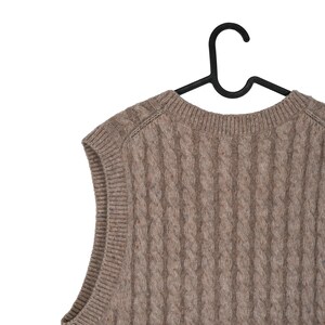 May include: A cable-knit, sleeveless sweater in a neutral brown color, hanging on a black hanger. The sweater has a textured pattern and a crew neck. The garment is made of a soft, possibly wool, material.