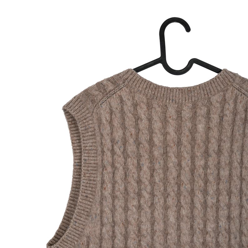 May include: A cable-knit, sleeveless sweater in a neutral brown color, hanging on a black hanger. The sweater has a textured pattern and a crew neck. The garment is made of a soft, possibly wool, material.