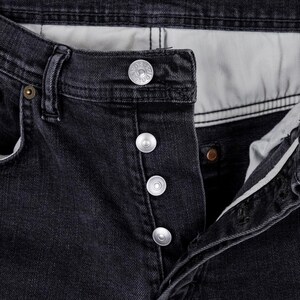 May include: Close-up of black denim jeans with silver button closure. The jeans have a classic five-pocket design and a button fly. The fabric appears to be slightly faded, giving the jeans a worn-in look.