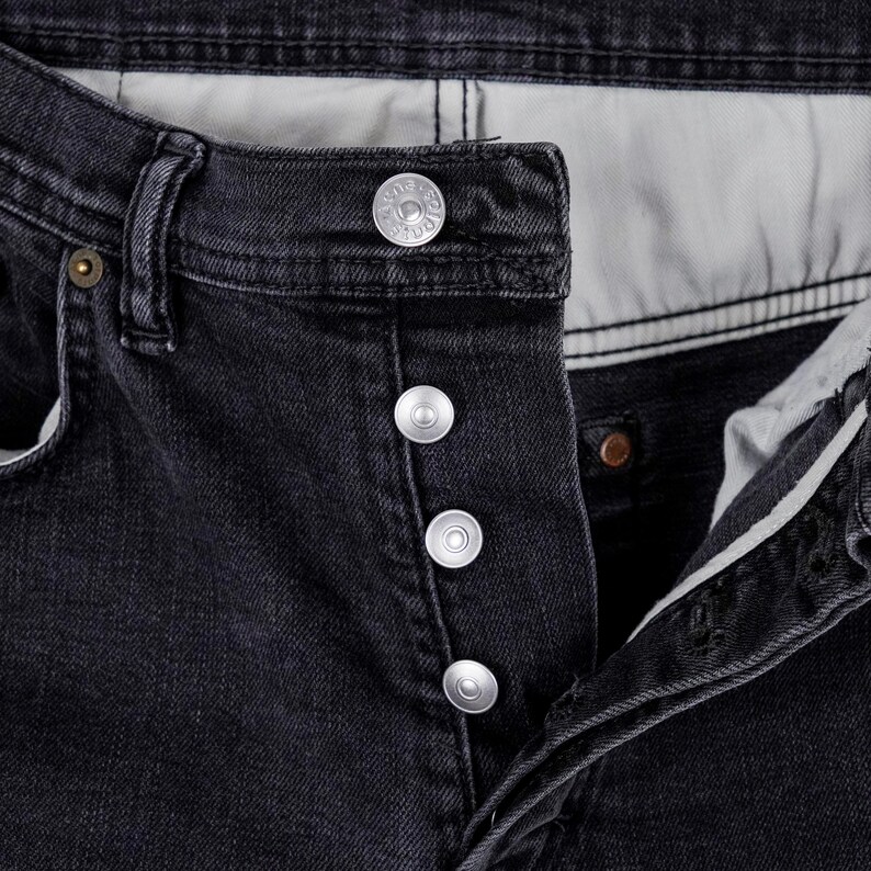 May include: Close-up of black denim jeans with silver button closure. The jeans have a classic five-pocket design and a button fly. The fabric appears to be slightly faded, giving the jeans a worn-in look.