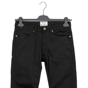 May include: Black denim jeans hanging on a black hanger. The jeans have a classic five-pocket design with a button and zipper closure. The brand name "Acne Studios" is visible on the hanger.