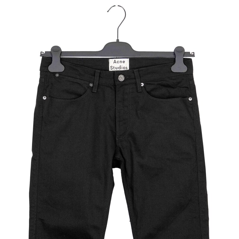 May include: Black denim jeans hanging on a black hanger. The jeans have a classic five-pocket design with a button and zipper closure. The brand name "Acne Studios" is visible on the hanger.