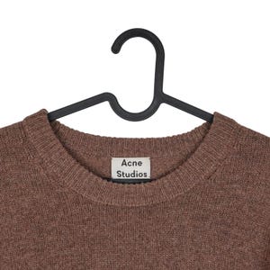 May include: A brown crew neck sweater hanging on a black hanger. The sweater has a ribbed neckline and the label reads "Acne Studios". The knitwear is a solid color and appears to be made of a soft material.