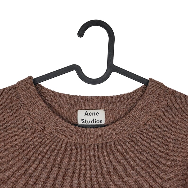 May include: A brown crew neck sweater hanging on a black hanger. The sweater has a ribbed neckline and the label reads "Acne Studios". The knitwear is a solid color and appears to be made of a soft material.