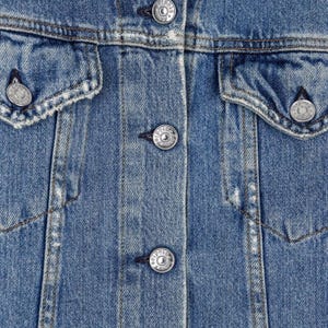 Op de afbeelding: Close-up van een lichtblauwe spijkerjas. De jas heeft zilverkleurige knopen, twee borstzakken en een klassieke kraag. De stof heeft een licht versleten look met zichtbare stikseldetails.