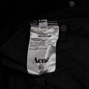 May include: Close-up of a black pair of jeans with a tag. The tag reads "MAX STAY CASH" and includes the brand name "Acne". The jeans are size 30/34. The tag also lists the materials and origin of the jeans.