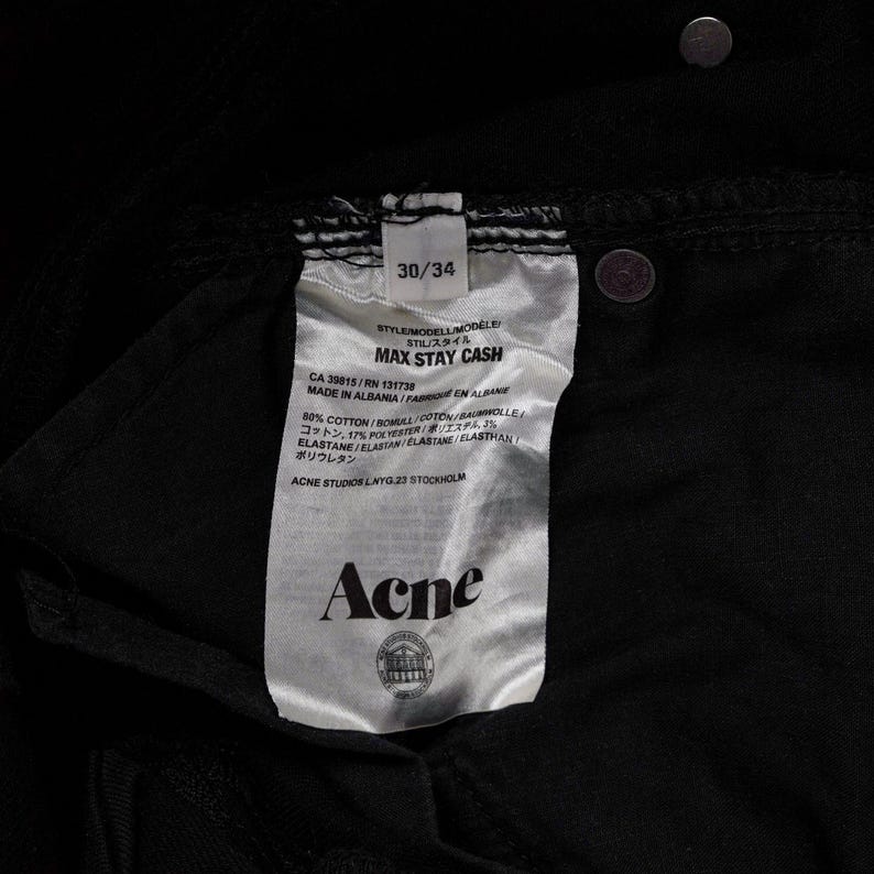 May include: Close-up of a black pair of jeans with a tag. The tag reads "MAX STAY CASH" and includes the brand name "Acne". The jeans are size 30/34. The tag also lists the materials and origin of the jeans.