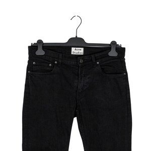 May include: Black denim jeans hanging on a black hanger. The jeans have a classic five-pocket design with a button and zipper closure. The brand name "Acne Studios" is visible on the hanger.