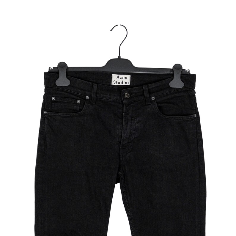May include: Black denim jeans hanging on a black hanger. The jeans have a classic five-pocket design with a button and zipper closure. The brand name "Acne Studios" is visible on the hanger.