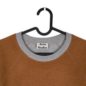 May include: A brown fleece sweatshirt with a grey crew neck and cuffs, hanging on a black hanger. The label reads "Acne Studios". The sweatshirt has a soft, textured appearance.