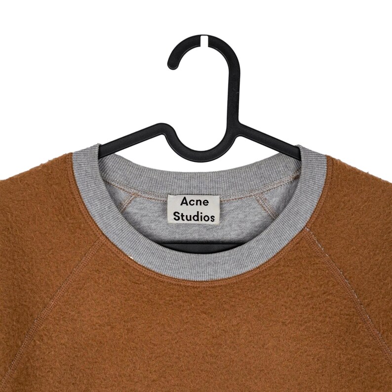 May include: A brown fleece sweatshirt with a grey crew neck and cuffs, hanging on a black hanger. The label reads "Acne Studios". The sweatshirt has a soft, textured appearance.