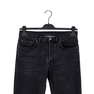 May include: Black denim jeans hanging on a black hanger. The jeans have a classic five-pocket design with a button and zipper closure. The denim appears to be a dark wash, with subtle fading and wear details.