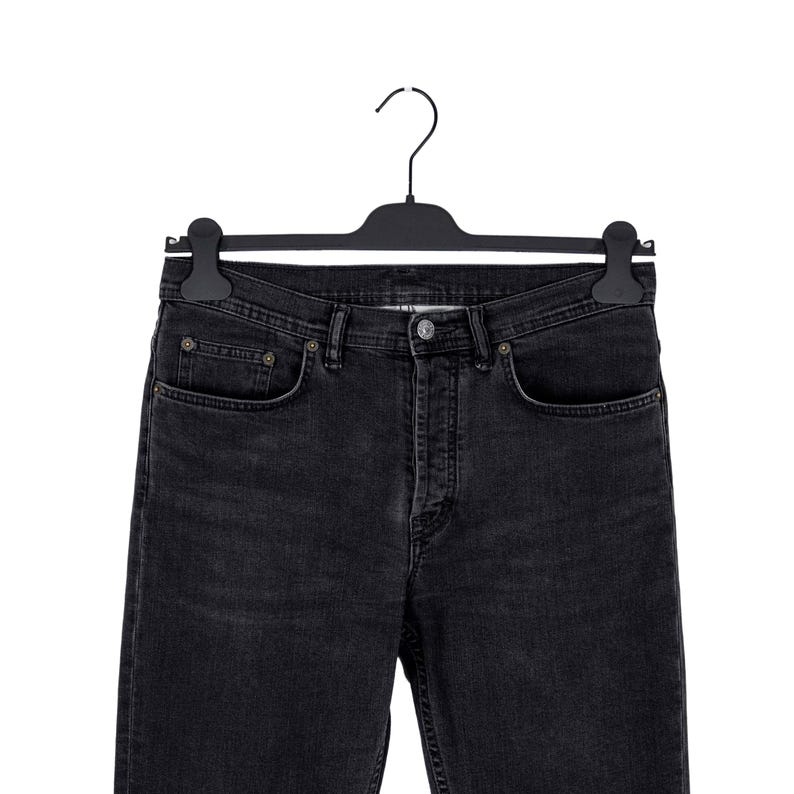 May include: Black denim jeans hanging on a black hanger. The jeans have a classic five-pocket design with a button and zipper closure. The denim appears to be a dark wash, with subtle fading and wear details.