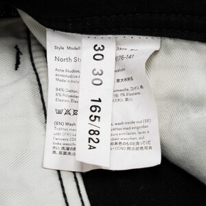 May include: A close-up of black jeans with a white label showing size 30/30 and brand details. The label indicates the jeans are from "Acne Studios" and made in Italy. Fabric composition details are also visible.